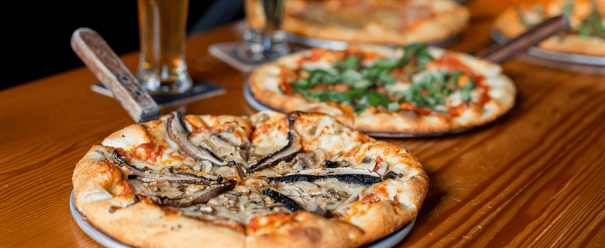 Four tasty lookig pizzas served with beer on a wooden restaurant table.